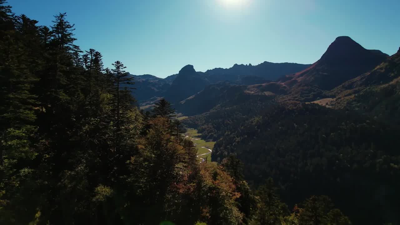 Wooded valley beneath rocky peaks backlit by the sun, dark fir trees in the foreground