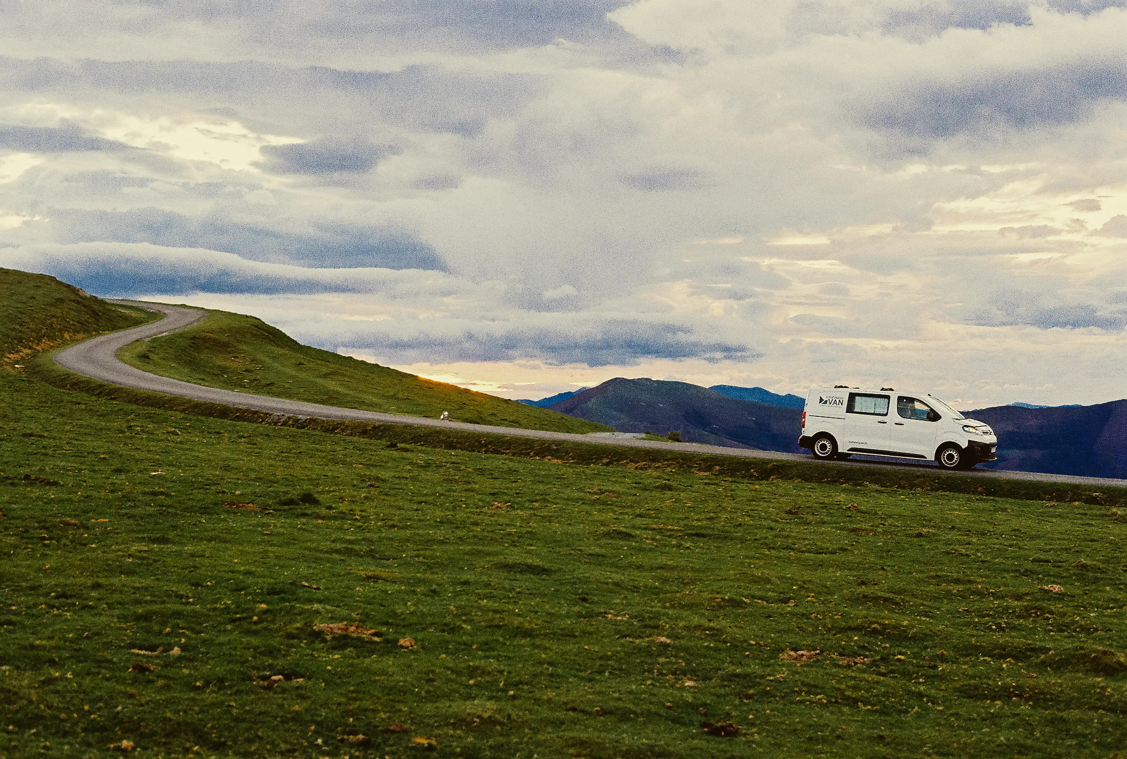 A white campervan on a grassy ridge road with low clouds hanging above the mountains