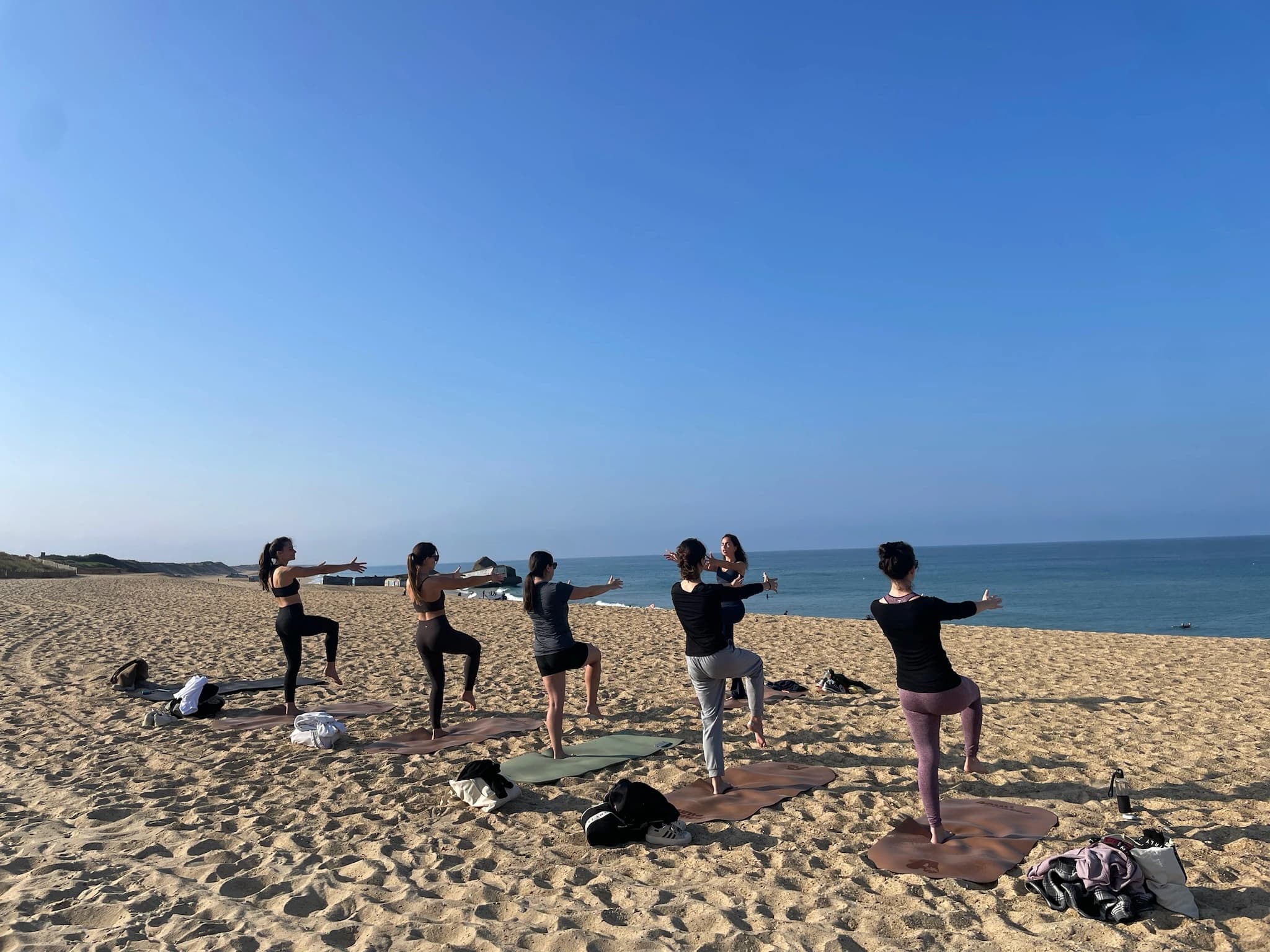 Five participants balancing with arms outstretched on mats facing the ocean on an empty beach