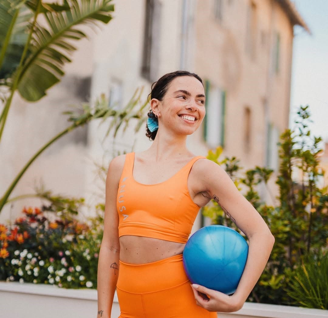 Smiling coach in orange sportswear holding a blue Pilates ball in a sunny courtyard