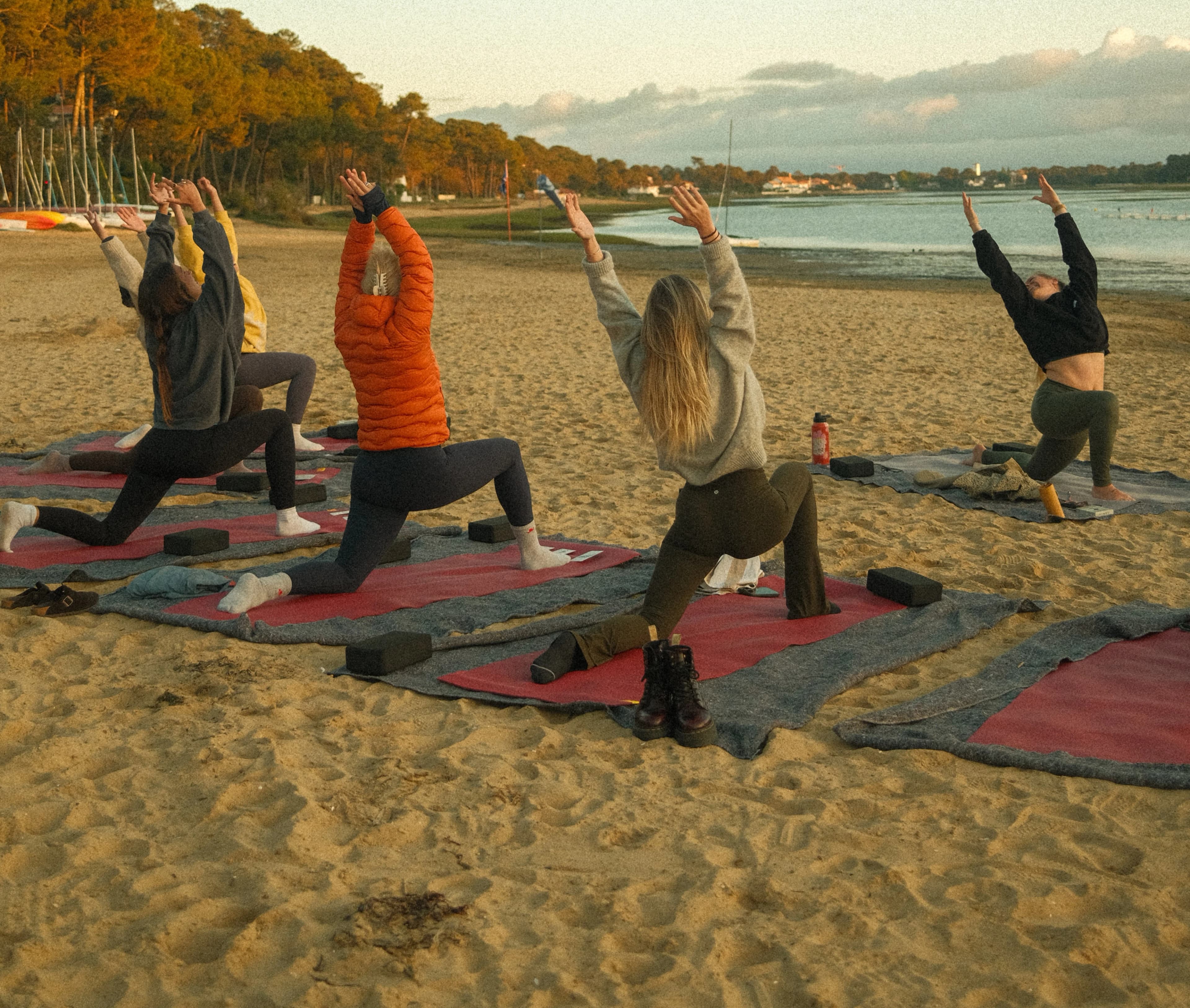 Group yoga class on the sand at sunset, low lunges with arms reaching up to the sky