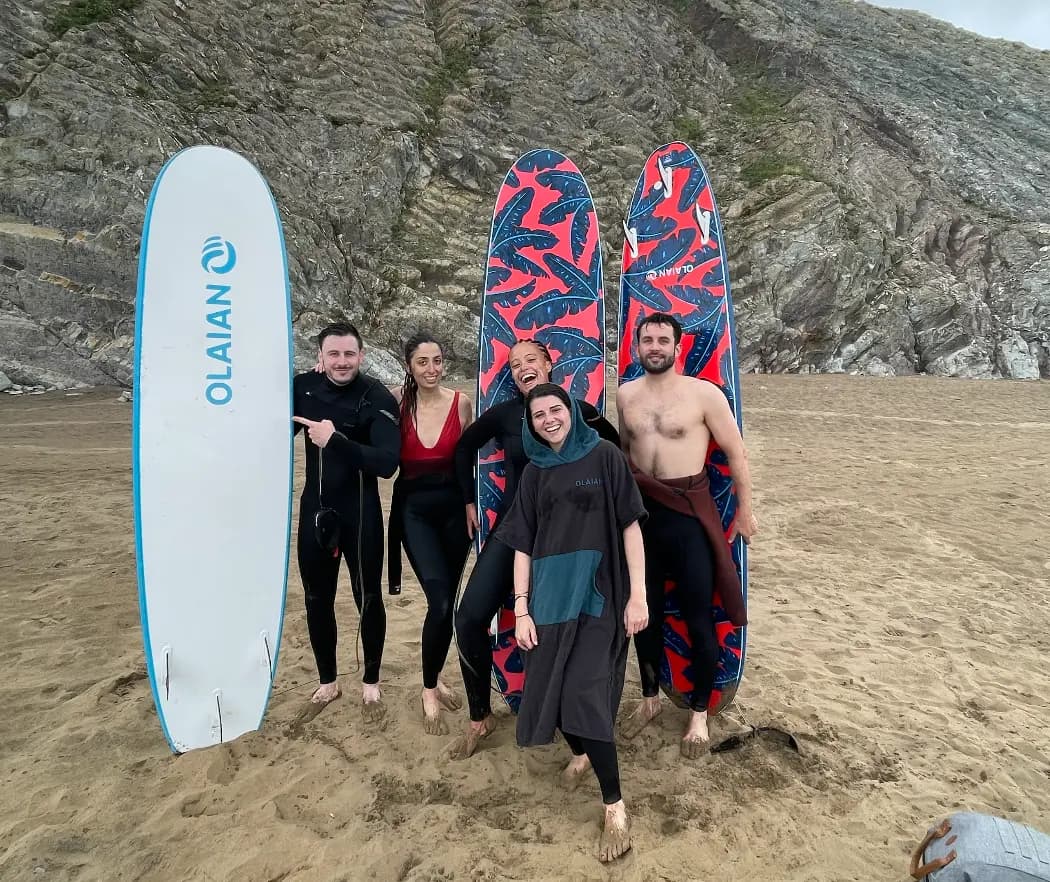 Five surfers smiling with their boards at the foot of a rocky cliff after a session