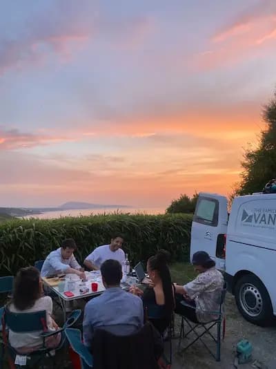 Group dinner in front of a campervan under a pink dusk sky, the coastline on the horizon