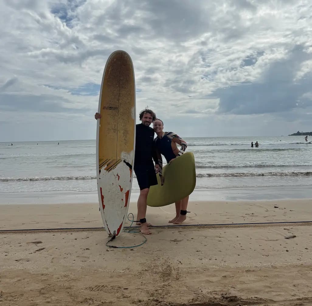 Two surf guides on the beach, one holding a longboard upright, the other a yellow board
