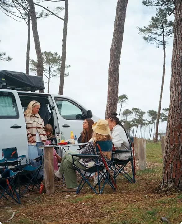 Group sitting at a table beside a campervan with its roof tent up, on a pitch among pines