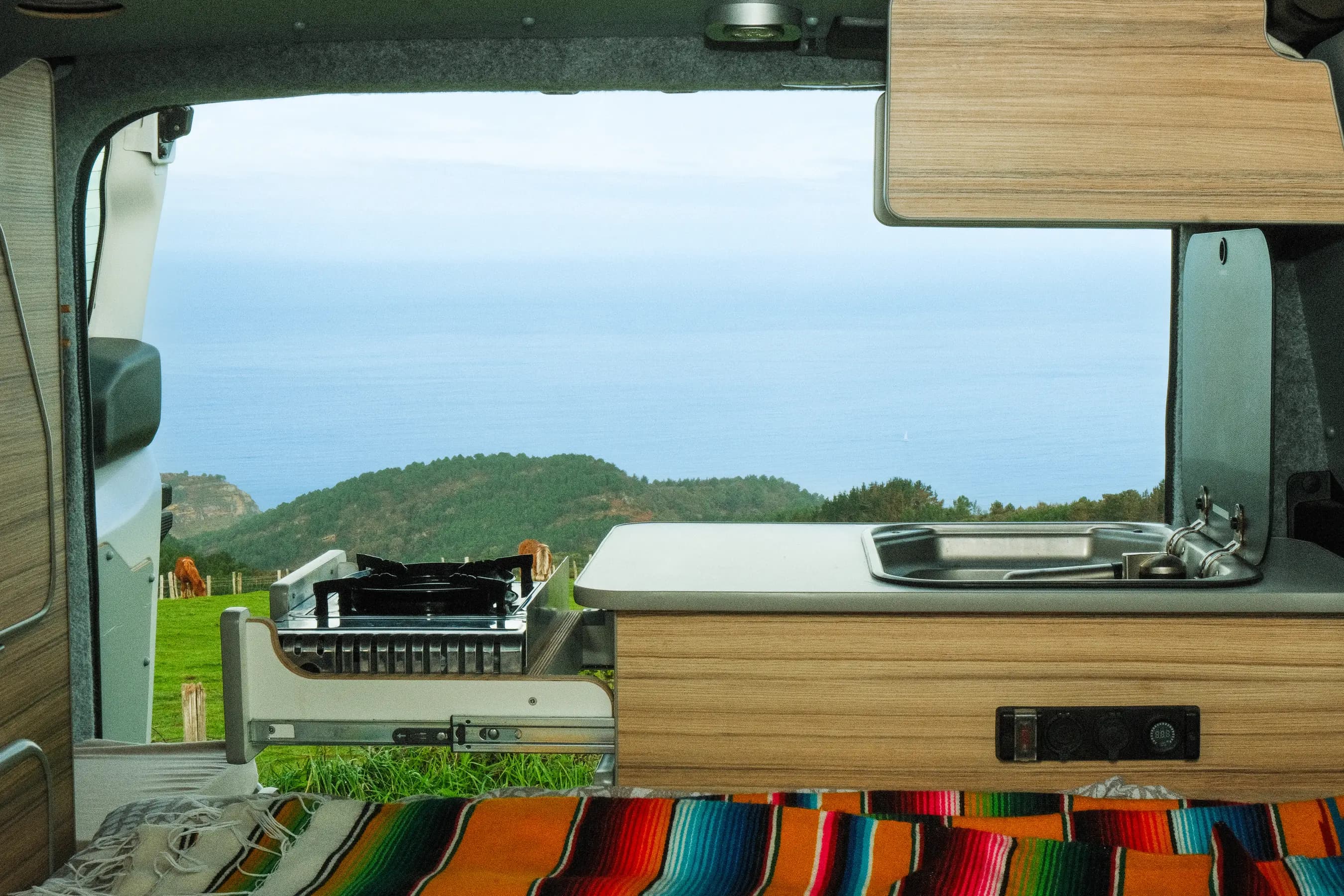 Campervan kitchen unit opening onto rolling hills and the sea, a striped blanket on the bed