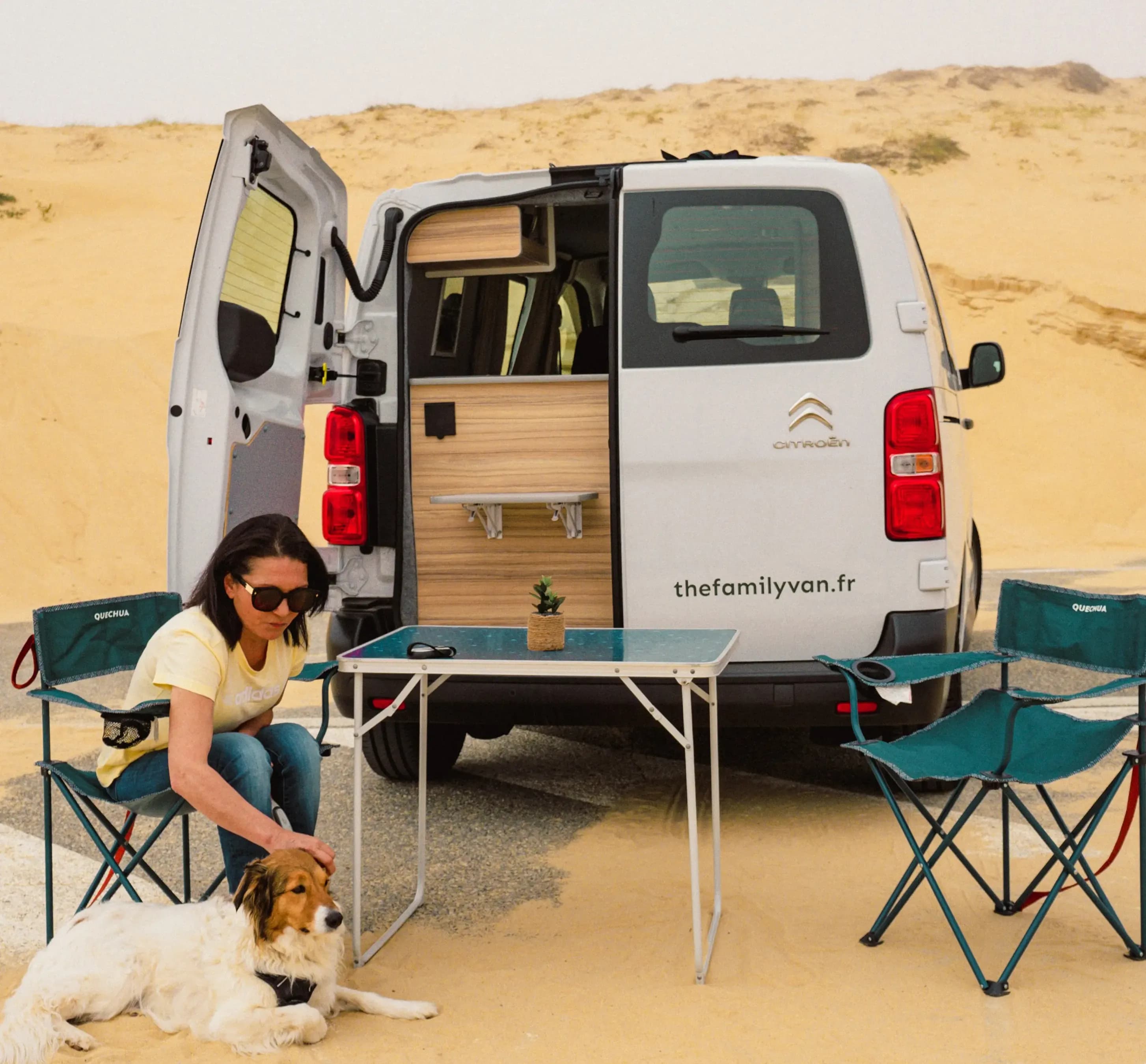 A woman stroking her dog beside a campervan with its rear doors open among the dunes