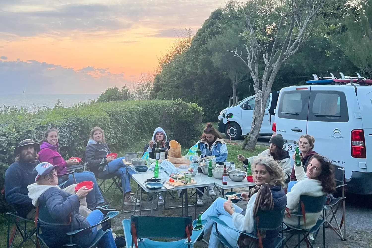 Shared camp dinner in front of the van, plates and bottles across two folding tables