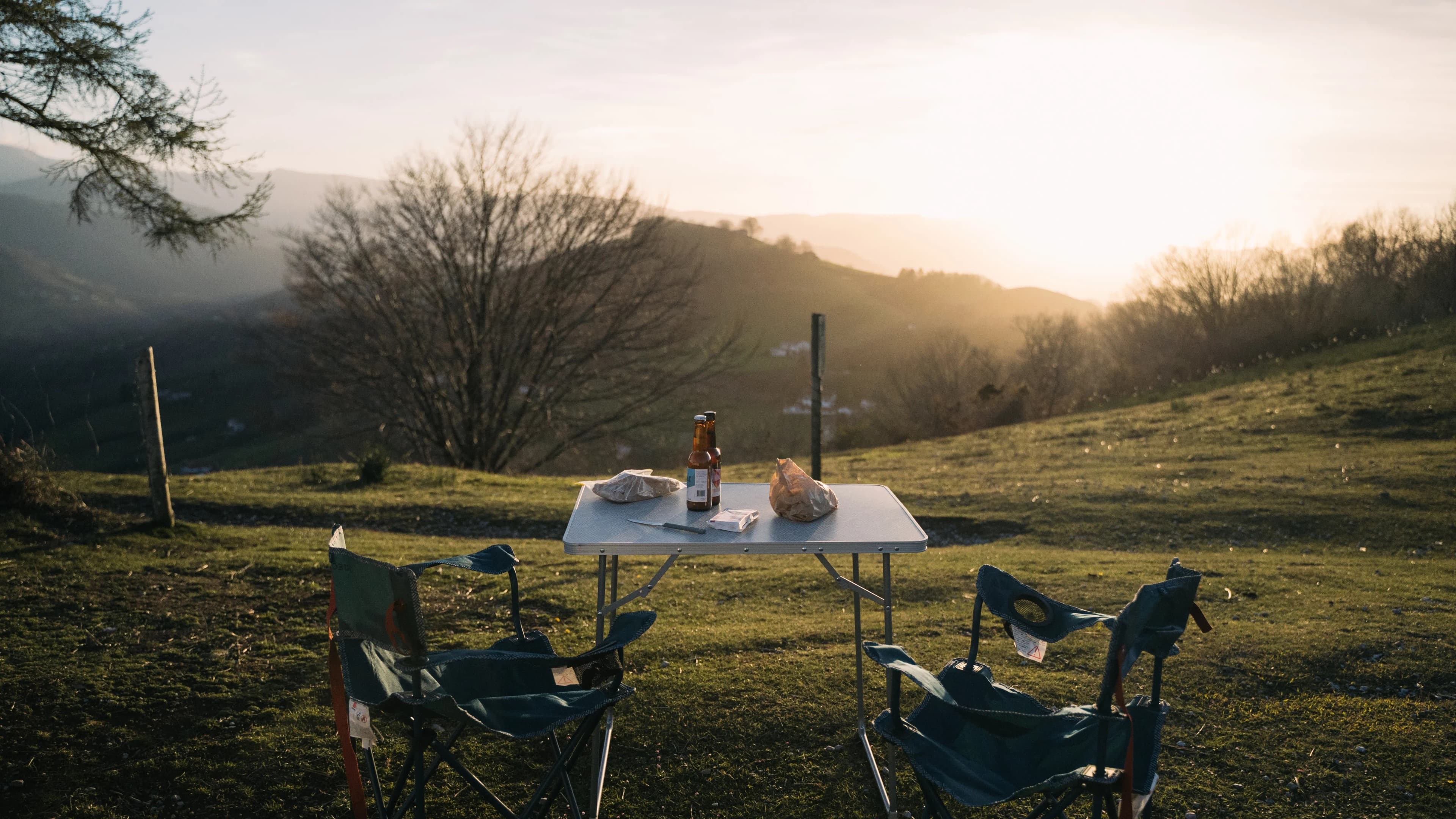 Camping table set with a beer, two folding chairs facing the hills in the evening light