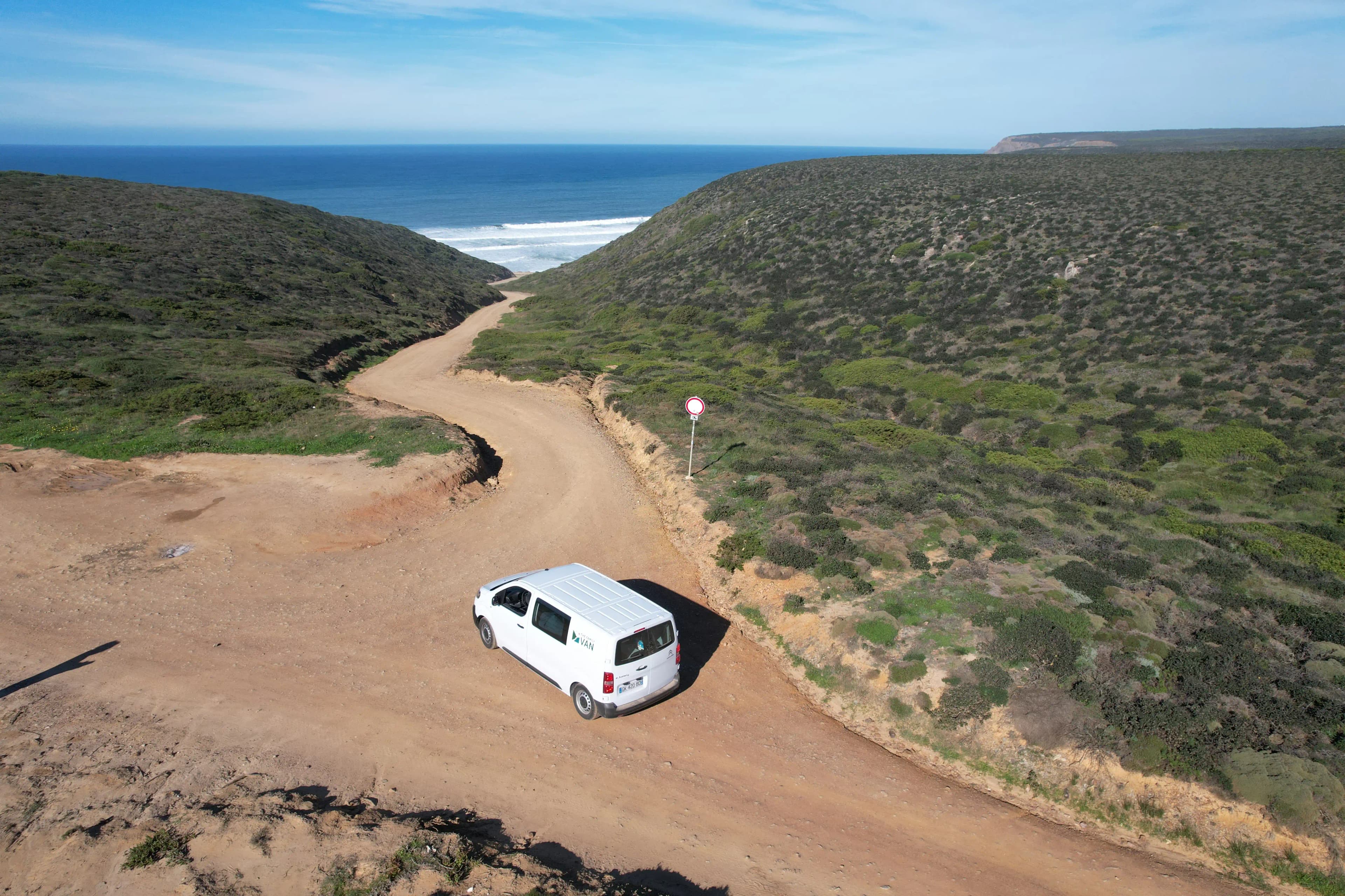 A white van on a dirt track winding down between green hills towards a surf beach