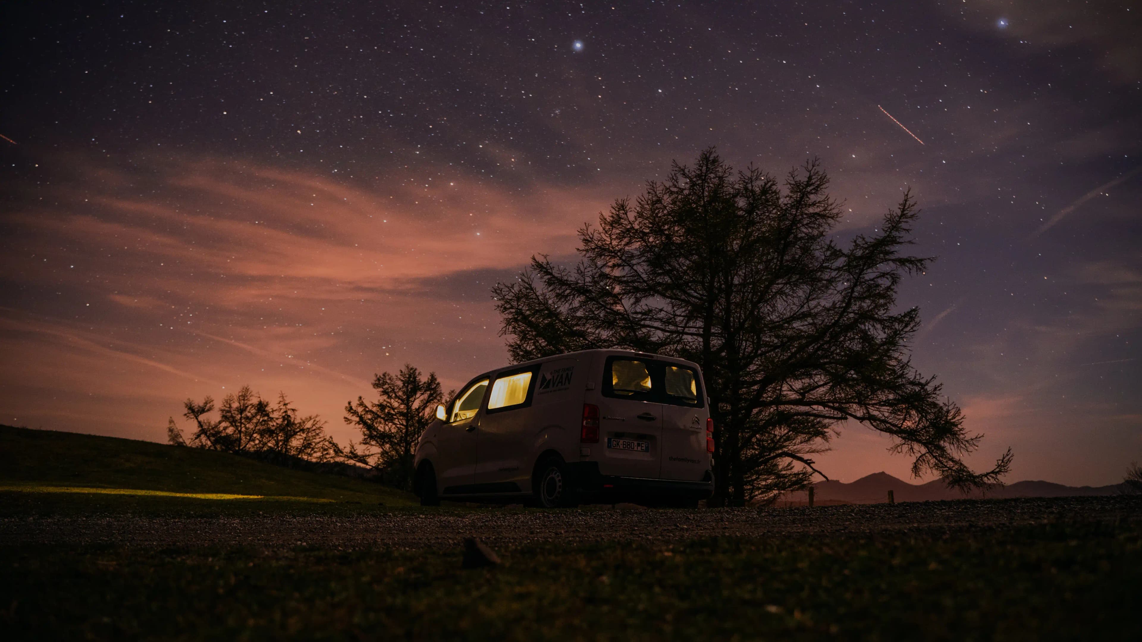 A campervan lit from within beneath a starry sky, a tree silhouetted above its roof