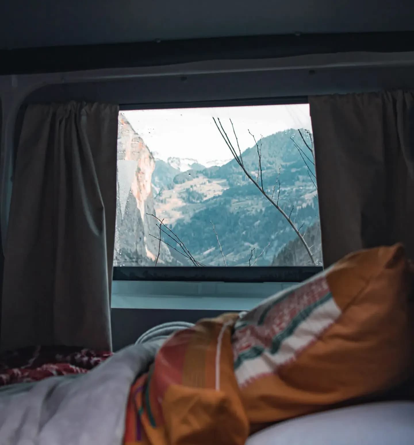 From the van's bed, the window frames a snow-dusted mountain valley above a colourful throw