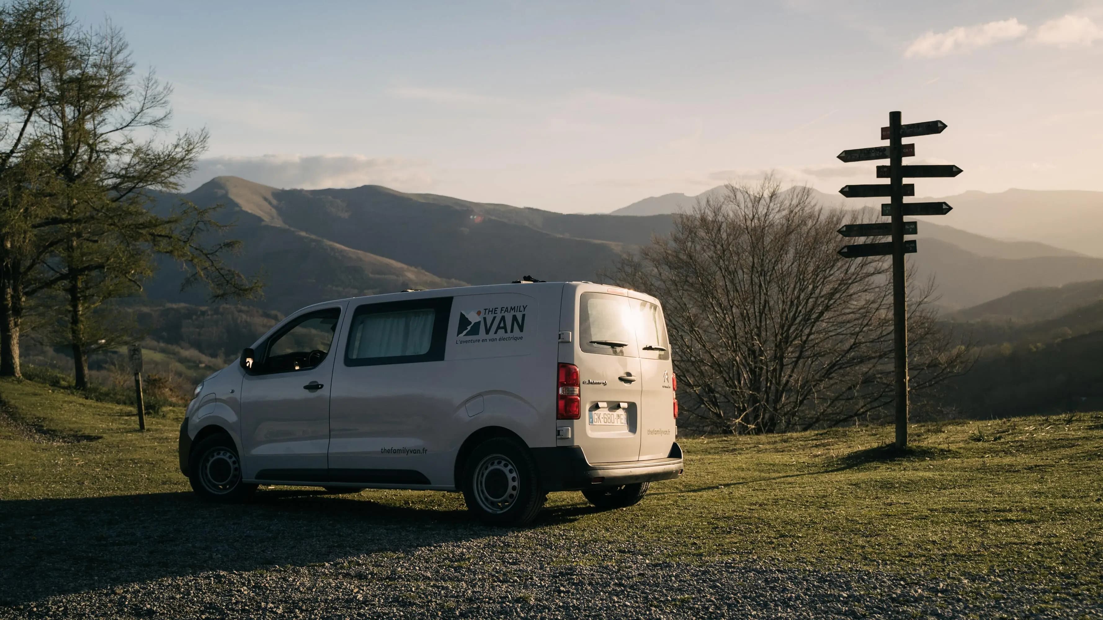 White campervan parked on a grassy pass beside a wooden signpost, mountains at sunset