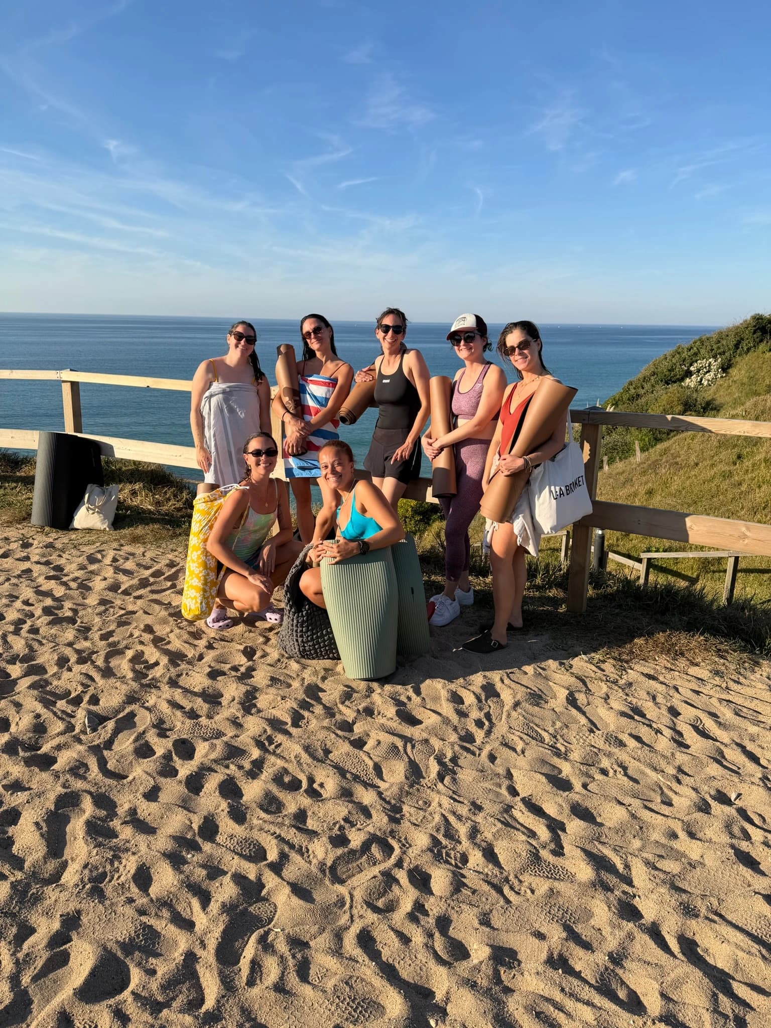 A group of women posing with their yoga mats on a sandy path high above the blue ocean