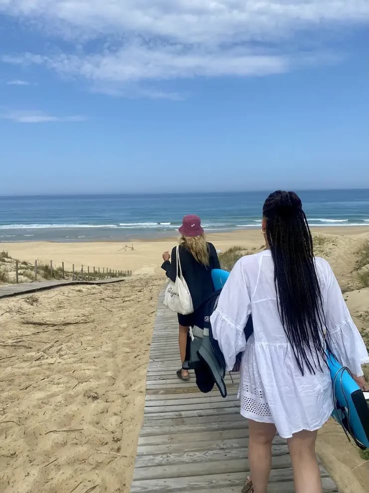 Two women walking down the wooden boardwalk to the beach, board and wetsuit in hand