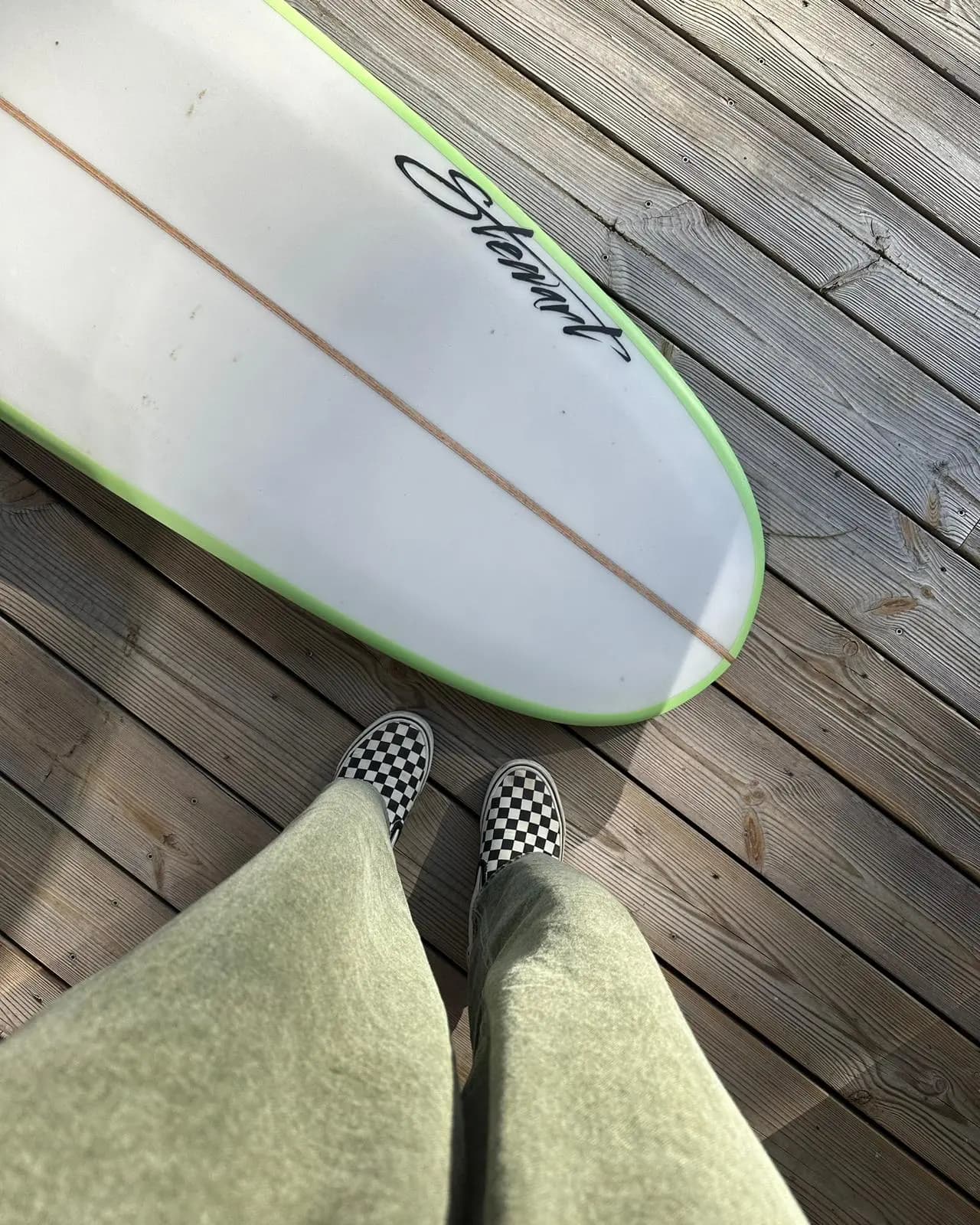 Surfboard resting on a wooden deck, legs and checked slip-on trainers in the foreground