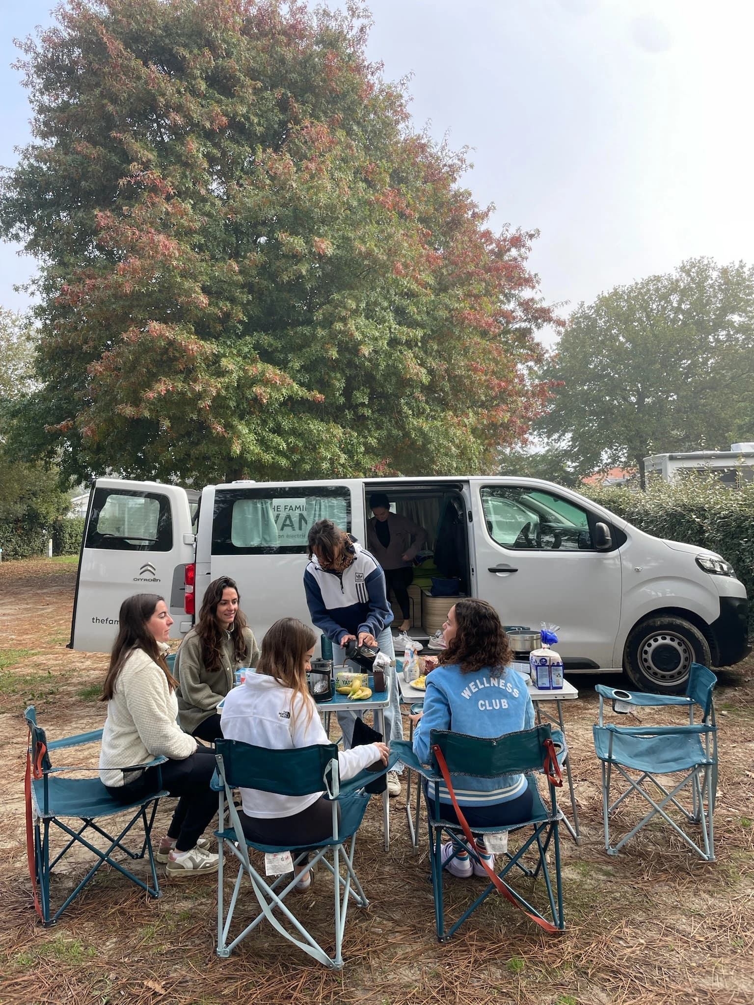 Women having breakfast at a camping table in front of a white van, autumn tree behind