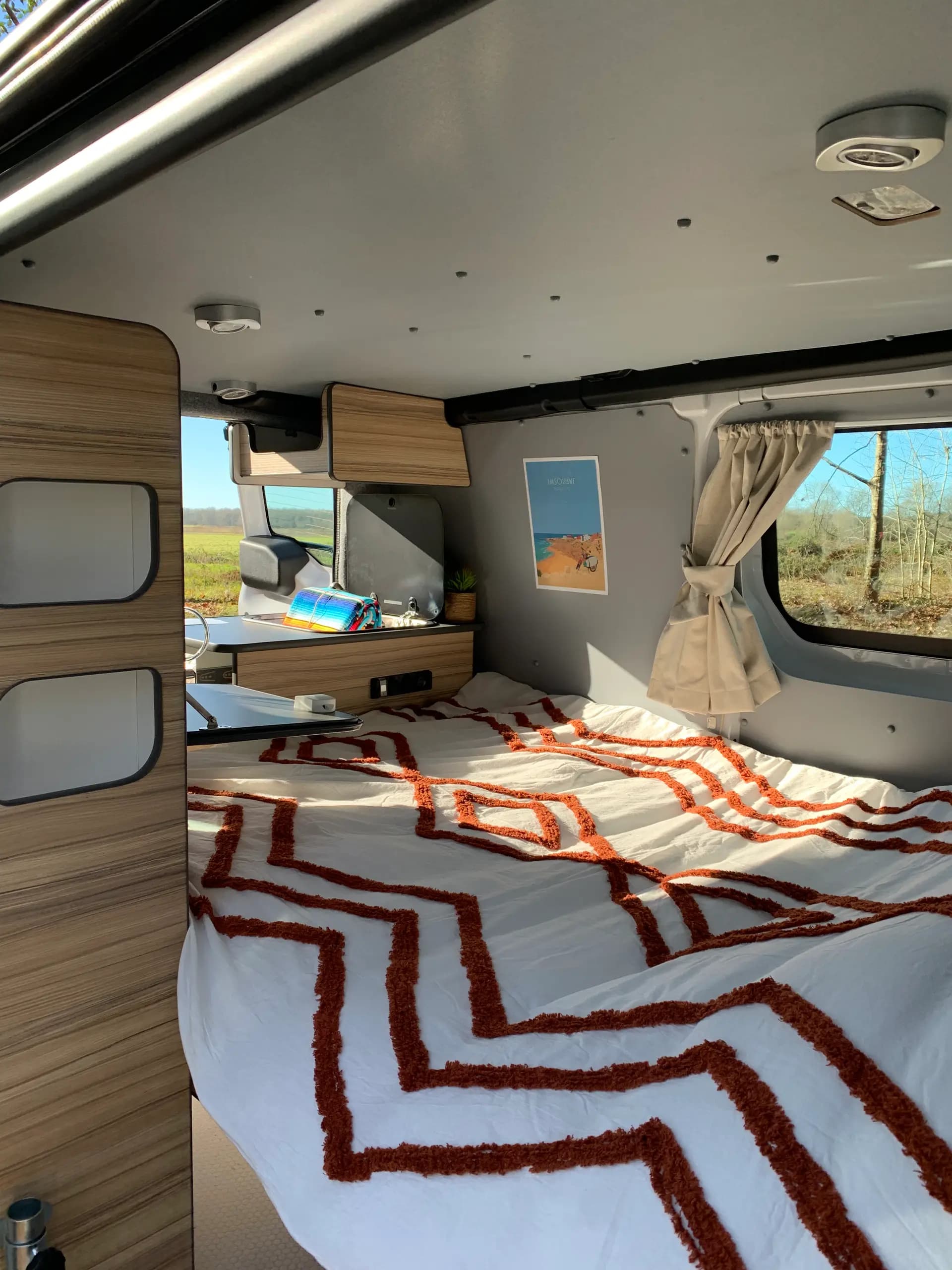 Double bed made up in a campervan, white bedspread with orange patterns and curtains at the window